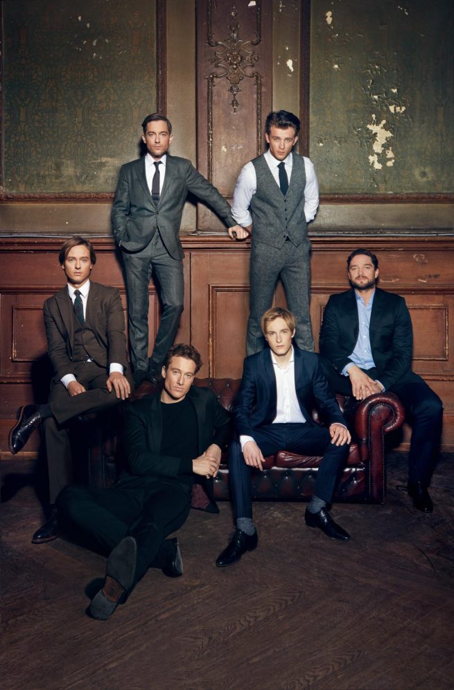 sechs prominente Schauspieler sitzen auf einer couch vor einer antiken wand mut Stuck. Sie tragen anzüge und hemden. sind elegant gekleidet.sie schauen ernst. manche lächeln ein bisschen. der boden ist aus holz und dunkel.